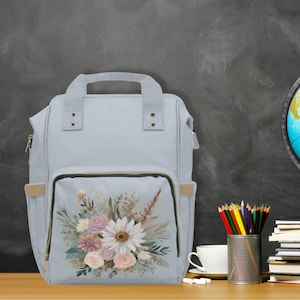 Puede incluir: Una mochila de lona azul claro con un estampado floral en el bolsillo delantero. El estampado presenta flores blancas, rosas y amarillas con hojas verdes.