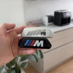 May include: Two car emblem badges. One is white with a gray and black logo, and the other is black with a blue, white, and red logo. The badges are held in a hand, with a blurred background.