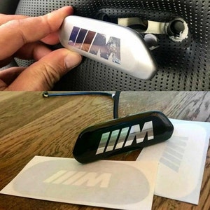 May include: A black and silver car emblem with the letters "M" in a stylised font. The emblem is on a black background and is surrounded by a silver trim.