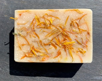 May include: A rectangular bar of handmade soap, pale beige in colour, decorated with bright orange and yellow flower petals. The soap has a speckled texture and a natural appearance, ideal for skincare, approximately 10cm long.