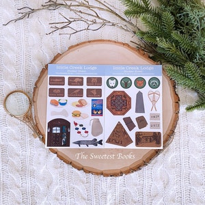 May include: Two sticker sheets featuring illustrations of animals, food, and geometric designs. The text "Icicle Creek Lodge" is displayed. The stickers are presented on a wooden slice, set against a white knitted background.