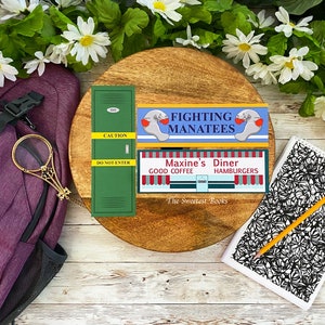 May include: A wooden round tray with three bookmarks. The bookmarks are green, blue and yellow with the text "Fighting Manatees", "Maxine's Diner Good Coffee Hamburgers" and "The Sweetest Books".