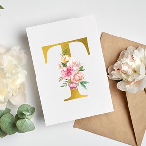 Pu&ograve; includere: Una lettera T dorata con un'arrangiamento floreale di fiori rosa, bianchi e gialli. I fiori sono disposti attorno alla lettera e sono circondati da foglie verdi. La lettera e i fiori sono su una carta bianca.