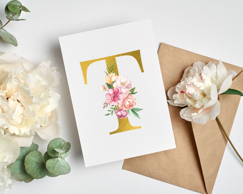 Pu&ograve; includere: Una lettera T dorata con un'arrangiamento floreale di fiori rosa, bianchi e gialli. I fiori sono disposti attorno alla lettera e sono circondati da foglie verdi. La lettera e i fiori sono su una carta bianca.