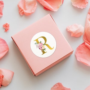 Pu&ograve; includere: Una scatola regalo rosa con un adesivo circolare bianco che presenta una lettera dorata "R" con accenti floreali.