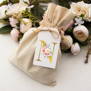 Pu&ograve; includere: Un piccolo sacchetto di tessuto beige con chiusura a cordoncino. Un'etichetta bianca con una lettera N dorata e un disegno floreale &egrave; attaccata al sacchetto. Il sacchetto &egrave; circondato da un bouquet di fiori artificiali bianchi e rosa.