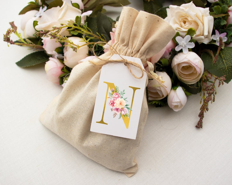 Pu&ograve; includere: Un piccolo sacchetto di tessuto beige con chiusura a cordoncino. Un'etichetta bianca con una lettera N dorata e un disegno floreale &egrave; attaccata al sacchetto. Il sacchetto &egrave; circondato da un bouquet di fiori artificiali bianchi e rosa.