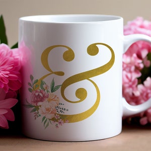Pu&ograve; includere: Tazza in ceramica bianca con un simbolo ampersand dorato e un design floreale. La tazza ha un manico ed &egrave; circondata da fiori rosa.