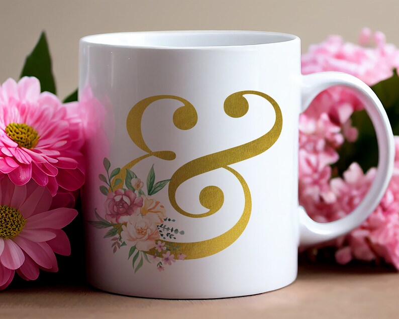 Pu&ograve; includere: Tazza in ceramica bianca con un simbolo ampersand dorato e un design floreale. La tazza ha un manico ed &egrave; circondata da fiori rosa.