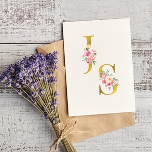 Pu&ograve; includere: Un biglietto bianco con una "J" e una "S" dorate con accenti floreali rosa e bianchi. Il biglietto &egrave; su una busta marrone e un mazzo di lavanda &egrave; sullo sfondo.