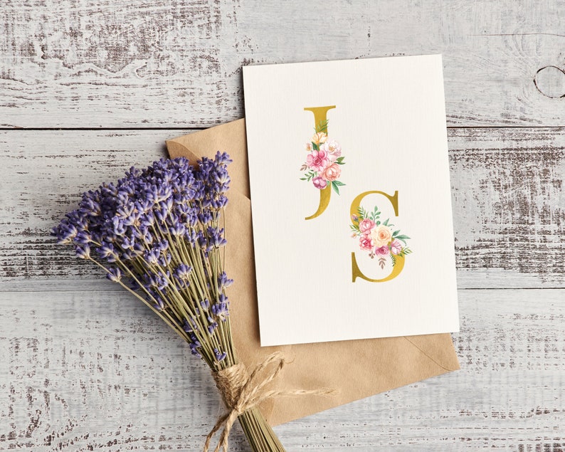 Pu&ograve; includere: Un biglietto bianco con una "J" e una "S" dorate con accenti floreali rosa e bianchi. Il biglietto &egrave; su una busta marrone e un mazzo di lavanda &egrave; sullo sfondo.