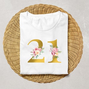 Pu&ograve; includere: Maglietta bianca con un numero 21 dorato decorato con fiori rosa e bianchi. Il numero &egrave; centrato sulla maglietta.