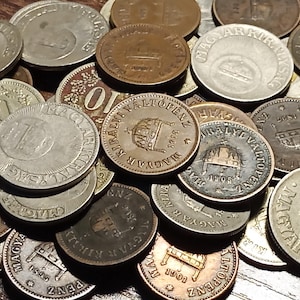 May include: A collection of vintage Hungarian coins, featuring various denominations and designs. The coins are made of silver and copper, and some have a gold-colored finish. The coins are arranged in a pile, with some overlapping.