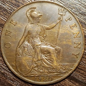 May include: A bronze-colored One Penny coin from 1920. The coin features a seated Britannia figure holding a trident and shield. The words "ONE PENNY" are inscribed around the edge.