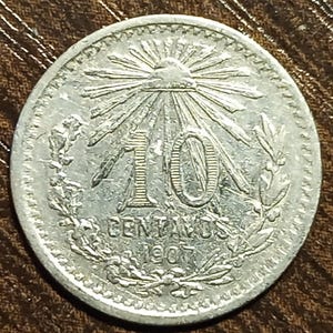 May include: A silver coin featuring the number "10" in the center, with the words "CENTAVOS" and the year "1907" below. The coin has a sunburst design and is encircled by a laurel wreath.