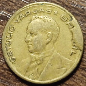May include: A gold-colored coin featuring a profile of a man in a suit and tie. The coin has the inscription "GETULIO VARGAS BRASIL" around the edge. The coin appears to be old and worn.