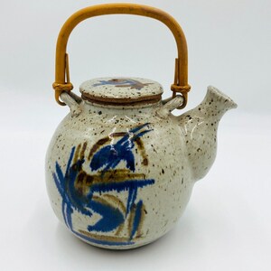 Può includere: Teiera in ceramica macchiata con manico in bambù. La teiera presenta disegni astratti blu e marroni. Il coperchio ha un design abbinato. Il beccuccio è sul lato destro della teiera.