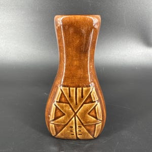 May include: A brown ceramic vase with a glossy finish. The vase has a geometric design in a lighter shade of brown on the lower section. The vase has a narrow neck and a wider base, suitable for displaying flowers or as a decorative item.