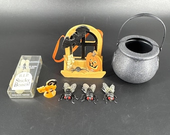 May include: A selection of Halloween decorations, including a black cauldron, a window with a pumpkin and bat, plastic flies, and a box of "Sticky Bones". The decorations are in various colours, including orange, black, and silver.