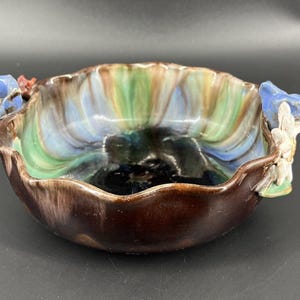 Puede incluir: Un cuenco decorativo de cerámica con un borde festoneado y un interior brillante multicolor. El cuenco presenta detalles florales azules y blancos a cada lado. El exterior es de color marrón oscuro.