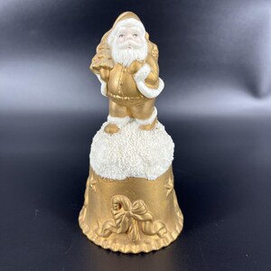 May include: A gold-colored ceramic bell featuring a Santa Claus figure standing on a snow-covered base. The bell has a detailed design with a bow and ribbon motif. The Santa figure is holding a Christmas tree.
