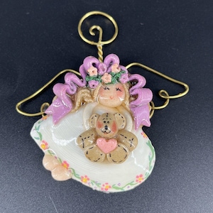 Puede incluir: Figura decorativa de ángel con un osito de peluche y un corazón. El ángel tiene un vestido blanco con detalles florales, un lazo rosa y un halo y alas doradas. El osito es marrón claro con un corazón rosa.