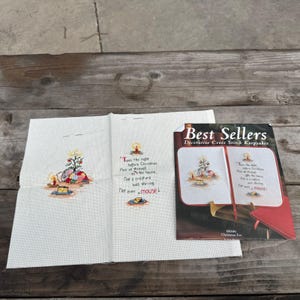 May include: A cross-stitch kit featuring a Christmas Eve design. The kit includes a piece of fabric with a stitched image of a Christmas tree, gifts, and candles, along with a booklet titled "Best Sellers". The text "'Twas the night before Christmas" is included.