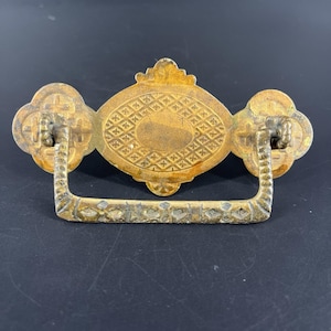 May include: Antique brass drawer pull with an ornate design. The pull features a central oval shape with a diamond pattern, flanked by two clover-shaped elements. A rectangular handle hangs below. The hardware is a warm, aged brass color.