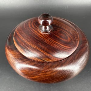 Op de afbeelding: Een ronde houten container met een deksel. De container en het deksel zijn gemaakt van donker, roodbruin hout met een opvallend nerfpatroon. Het deksel heeft een kleine, afgeronde knop voor gemakkelijk hanteren. De container wordt waarschijnlijk gebruikt voor opslag.