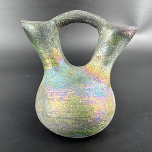 May include: A handcrafted, iridescent ceramic vase with a unique double-spout design and a circular handle. The vase features a pearlescent finish with hues of green, gold, and purple, creating a shimmering effect. The vase has a rounded base and a textured surface.
