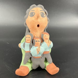 Puede incluir: Figurilla de cerámica hecha a mano que representa a una persona sosteniendo tres figuras más pequeñas. La figura principal tiene una expresión de sorpresa. Las figuras están pintadas en naranja, gris, verde y azul.