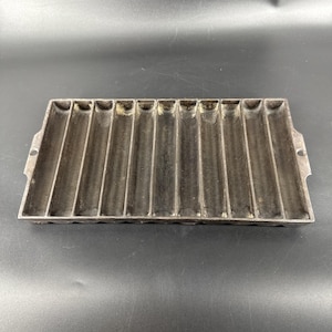 May include: A rectangular, cast iron cornbread pan with ten individual molds. The pan is a dark grey colour and has two small handles on either side. The molds are long and narrow, perfect for baking cornbread sticks.