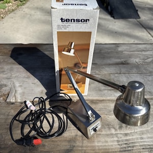 Może przedstawiać: Srebrna lampa biurkowa Tensor Mini Swivel z pudełkiem. Lampa ma obrotowe ramię i cylindryczną podstawę. Na pudełku widnieje napis "Tensor" i "High Intensity Lamp". Lampa stoi na drewnianej powierzchni.