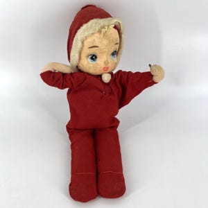 Puede incluir: Muñeca vintage con rostro pintado, ojos azules y atuendo rojo. La muñeca lleva una capucha roja con ribete color crema. Los brazos de la muñeca están extendidos y tiene un pequeño botón redondo en el pecho. El atuendo de la muñeca incluye una parte superior de manga larga y pantalones.