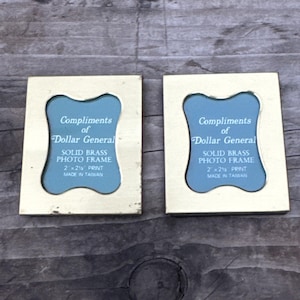 Puede incluir: Dos marcos de fotos rectangulares de latón macizo con acabado dorado. Cada marco presenta un recorte central con una inserción azul impresa con el texto: Compliments of Dollar General, SOLID BRASS PHOTO FRAME, 5,08 x 6,35 cm, MADE IN TAIWAN.