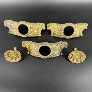 Op de afbeelding: Antieke messing hardware set. De set bevat drie decoratieve rozetten en twee bijpassende ladeknopen. De rozetten hebben een gebogen vorm met een centraal gat en gedetailleerde bloemen- en krulwerk. De ladeknopen hebben een bloemenontwerp.