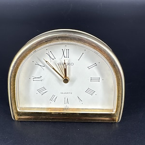 May include: A gold-coloured Seiko quartz clock with a white face and black Roman numerals. The clock has a semi-circular shape and features gold hands. The word "SEIKO" is printed on the face.