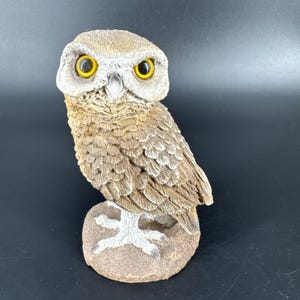 May include: A white ceramic mug with a black handle and the text "But First, Coffee" in a bold, black font. The mug is set against a blurred background, suggesting a cozy setting.