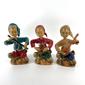 May include: Three vintage figurines of musicians. Each figure is dressed in colourful clothing and playing a different instrument: a flute, a basket, and a lute. The figurines have gold-coloured bases and painted faces.