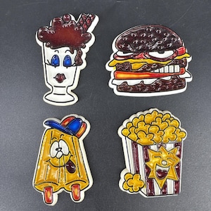 Puede incluir: Cuatro imanes coloridos con temática de comida, estilo caricatura. Incluye un helado de chocolate, una hamburguesa con queso, un personaje de paleta con gorra de béisbol y una caja de palomitas de maíz sonriente.