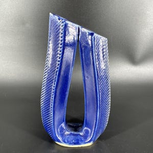 May include: A striking, cobalt blue ceramic vase with a unique, open design. The vase features a textured, ribbed pattern on the exterior, with a smooth, glossy finish. The form is tall and slender, with a distinctive, angled top.