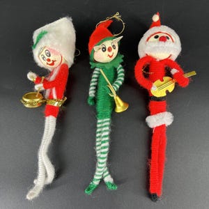 Può includere: Tre decorazioni natalizie vintage. Uno è un elfo rosso e bianco che suona un tamburo, un altro è un elfo verde e bianco con una tromba, e il terzo è un Babbo Natale che suona una piccola chitarra d'oro. Tutti sono fatti di materiale sfocato.