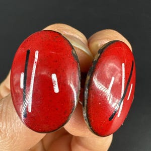 以下が含まれることがあります： 白と黒の線形デザインが施された、楕円形の赤いイヤリングのペア。イヤリングは光沢のある仕上げと暗い縁取りが特徴です。デザインは抽象的でモダンで、普段使いや特別な機会に適しています。