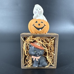 Puede incluir: Una decoración temática de Halloween con una calabaza naranja sonriente coronada por un fantasma blanco. Debajo, un gato negro con sombrero y pajarita está sentado en una caja de madera llena de paja. La exhibición tiene una apariencia rústica y artesanal.