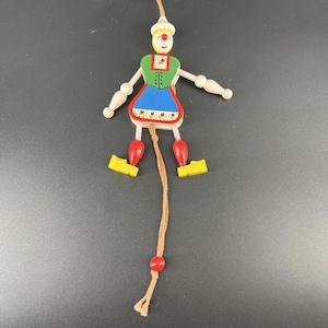 May include: A painted wooden pull-string puppet. The puppet features a white hat, green top, blue skirt, and red and yellow details. The arms and legs are jointed, and it is suspended by a string. The puppet is a traditional toy.
