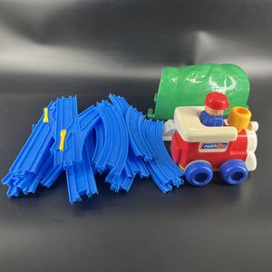 May include: A colorful toy train set featuring a red, white, and blue train with a driver figure, blue tracks, and a green tunnel. The train has the text "Push 'n Go" on the side. The set is designed for young children.