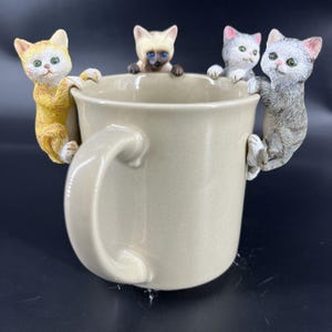 以下が含まれることがあります： ベージュ色のセラミックマグカップ。縁には4匹の猫の装飾品が付いています。猫は黄色、グレー、シャムなど様々な色で、詳細な特徴と緑色の目があります。マグカップには大きな取っ手が付いています。