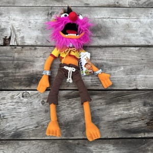 May include: A plush Animal character from The Muppets featuring a bright pink shaggy mane, a red nose, and a wide open mouth with white teeth. The figure wears a yellow shirt, brown pants with a white rope belt, and silver wrist cuffs.