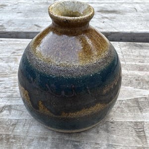 May include: A round ceramic vase with a narrow neck and a flared rim. The surface features a layered glaze with a glossy golden-brown top section that transitions into a deep, textured dark blue and brown mottled finish across the spherical body.