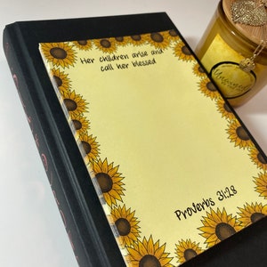 May include: A black book with a sunflower-decorated notepad on top. The notepad has the text "Her children arise and call her blessed" and "Proverbs 31:28". A candle and necklace are in the background.